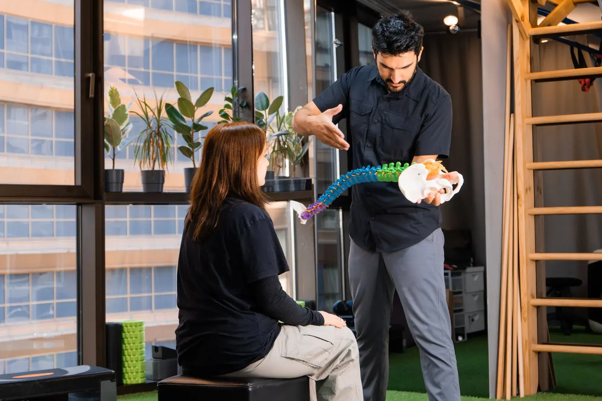 A practitioner holding a full spine and pelvis model out toward a seated woman, his free hand shaping the movement he is describing.