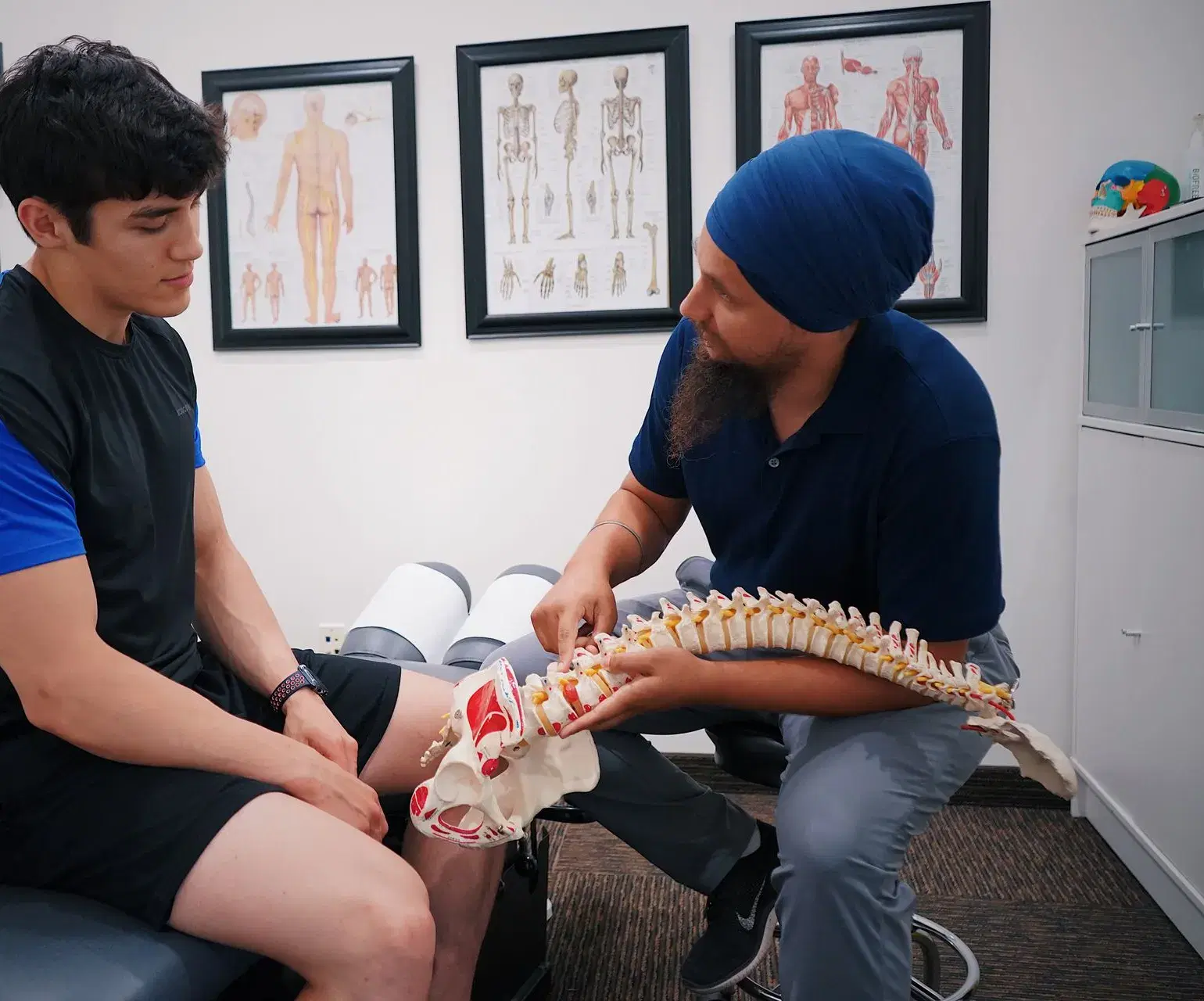 A seated practitioner turning a lumbar spine and pelvis model toward a patient, one finger resting on a single level.