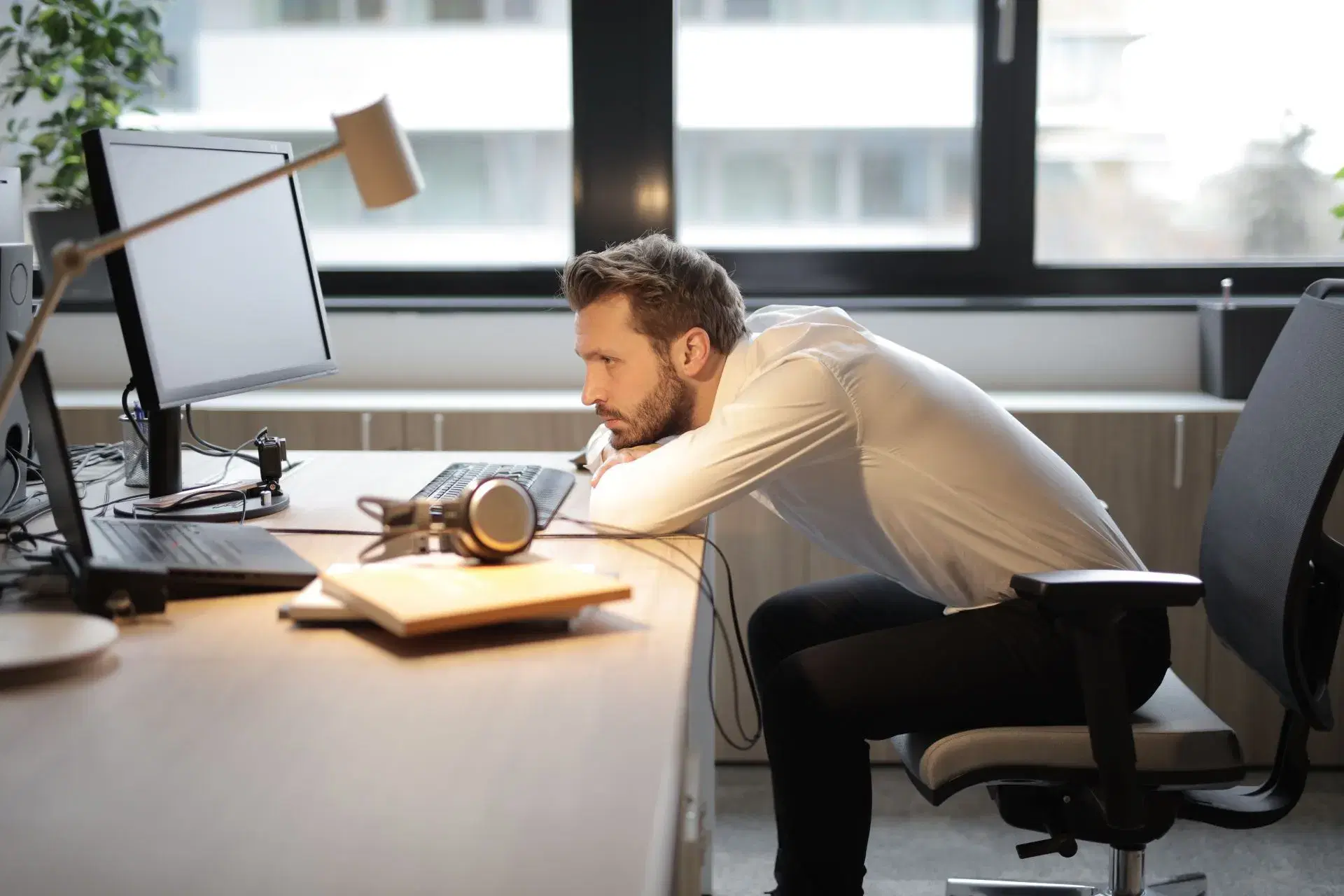 A man at a desk seen from the side, folded forward over the keyboard with his head craned toward the monitor.