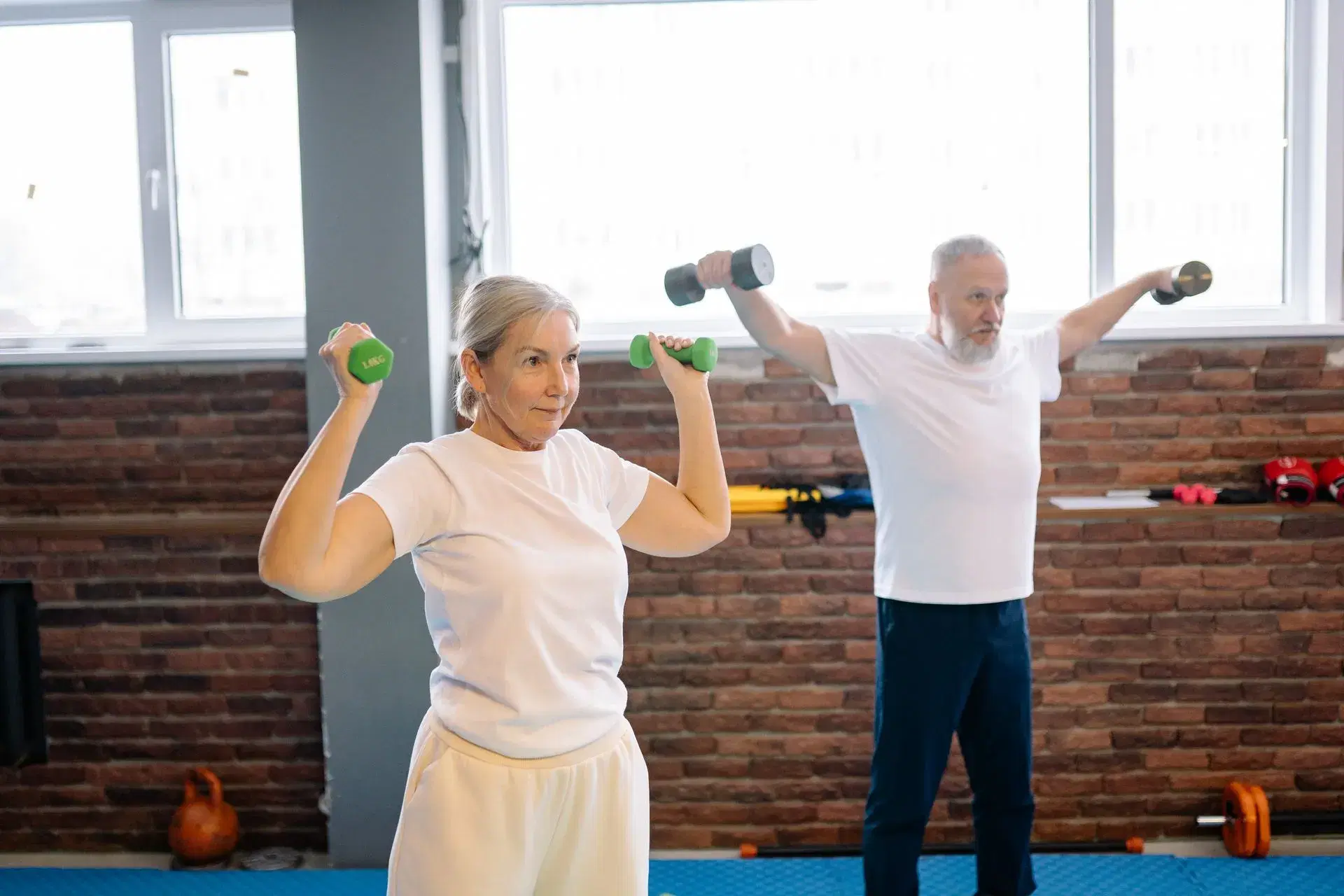 An older man and woman raising light dumbbells to shoulder height in a bright room with a brick wall behind them.