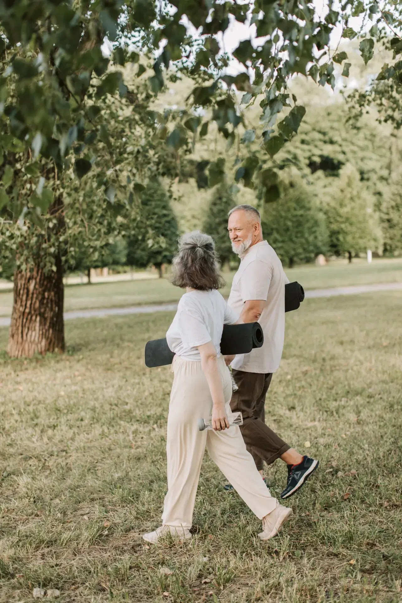 An older couple walking through a park carrying exercise mats.