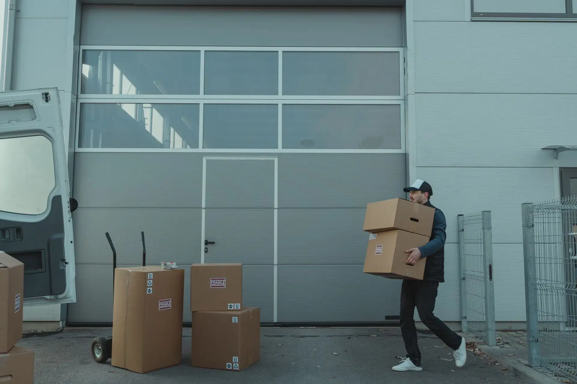 A delivery worker carrying two stacked cardboard boxes in close to the chest, walking from a van toward a roller door.