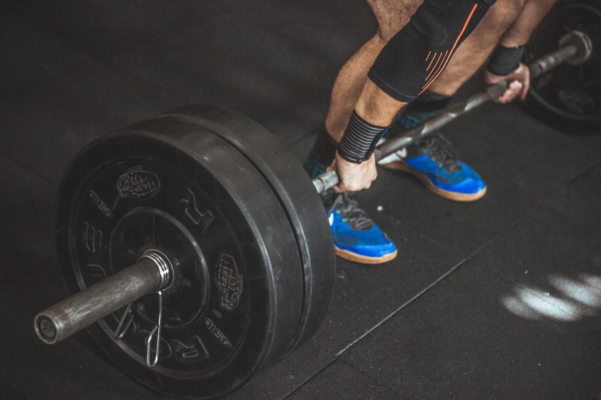 A waist-down view at the bottom of a deadlift setup: hands on the knurl, shins close to a loaded barbell.