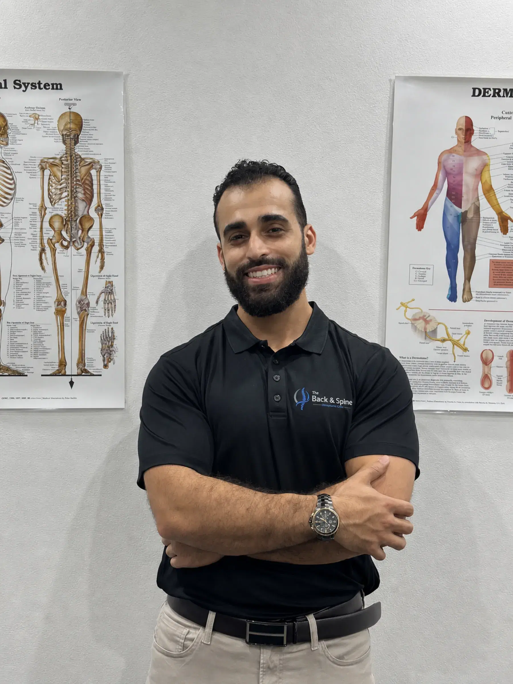 Dr Laith Musallam, chiropractor, arms crossed in front of the clinic's anatomy charts.