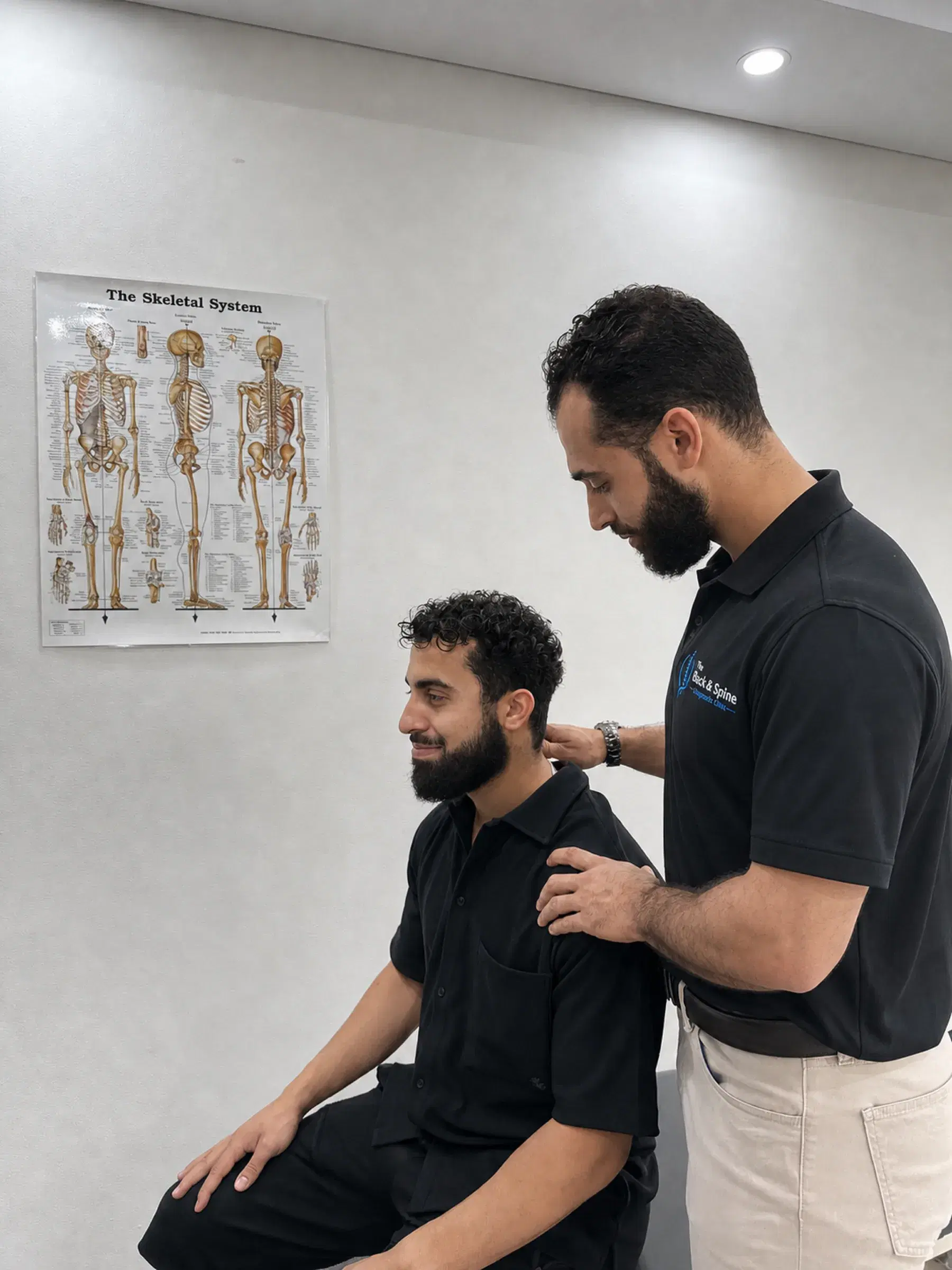 Dr Laith Musallam assessing the neck and shoulders of a seated patient, a skeletal-system chart on the wall behind them.