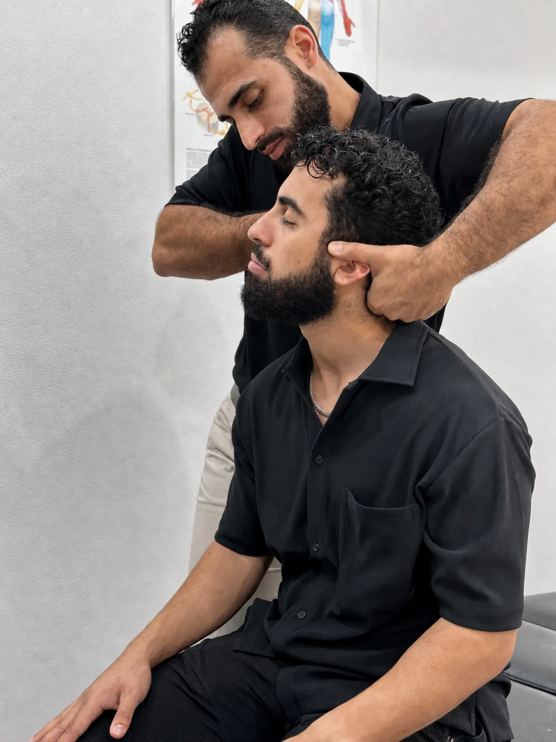 Chiropractor Dr Laith Musallam standing behind a seated patient, both hands cradling the head and neck, the patient's eyes closed.