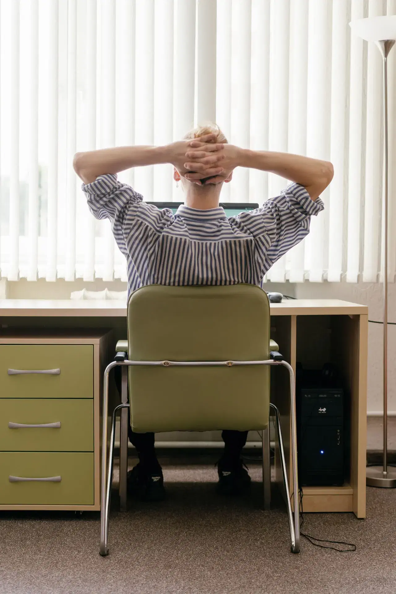 A person seen from behind at a desk, hands laced behind their head, window blinds ahead of them.